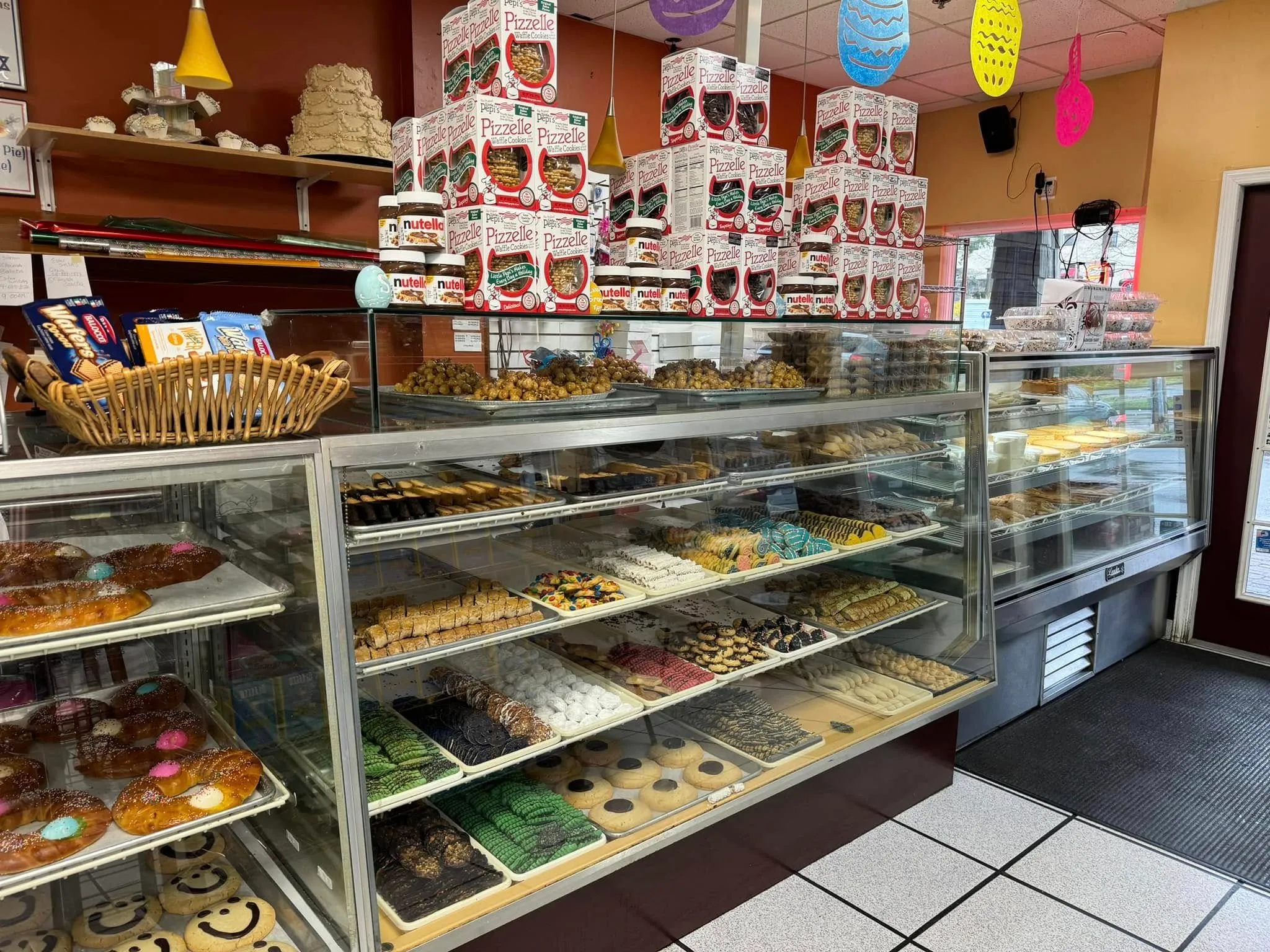 Glass bakery cases with trays of Italian cookies and Easter bread rings, between a terracotta wall and a mustard wall